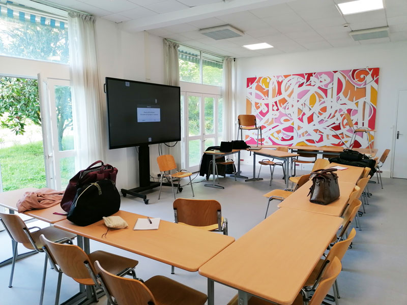 Une salle avec des tables, et un écran tactile. Au mur, une fresque.