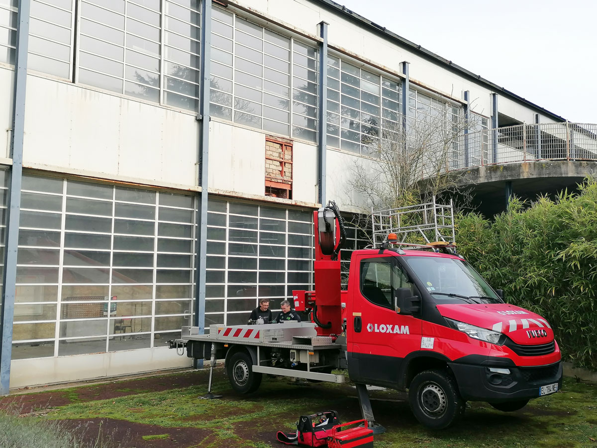 Une vue sur le bâtiment collectif. Devant un camion avec un bras élévateur permettant d'observer en hauteur la structure.